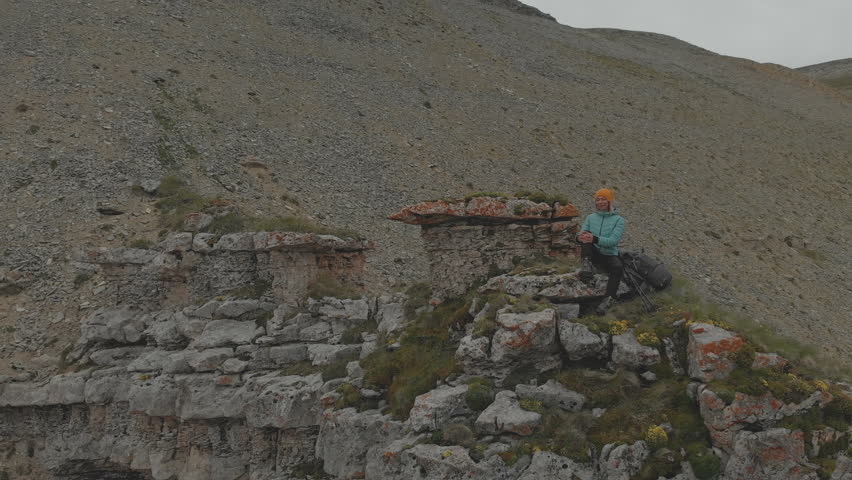 Aerial - flight around young adult woman with a backpack sits and rests of the rock at the foot of the epic plateau at sunset. View from the back. Russia. North Caucasus