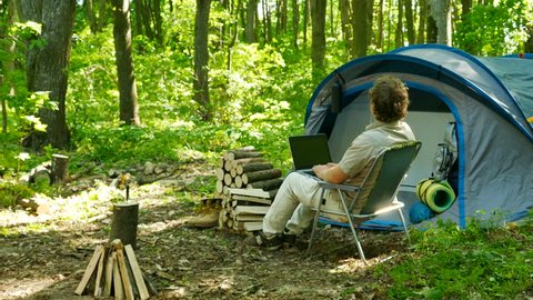 4k.Modern man tourist works notebook near tent in wood, Camping team