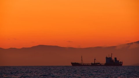 A huge cargo ship floatsin the sea in the yellow rays of the evening sun against the background of distant mountains