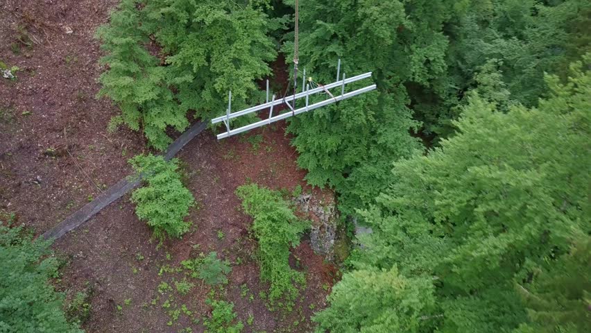 Metal bridge of pathway in forest is lifted by crane down into the gorge aerial drone shot working people