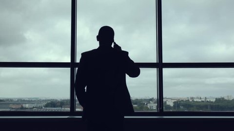 Silhouette of businessman in jacket near large panoramic window talking on the smartphone.The head approaches the window in office and starts telephone conversation. Important phone call for business.