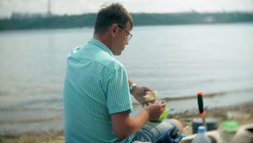 A man is eating around a kettle in a campsite with a tent on the background. 4k