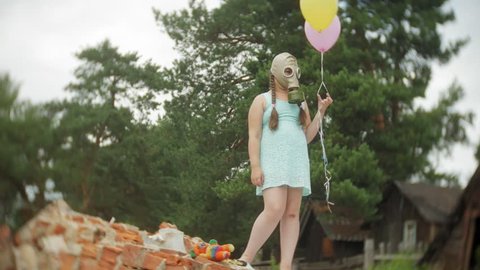 A little girl in a gas mask on the ruins of a building and holding on to a doll and balloons. 4k
