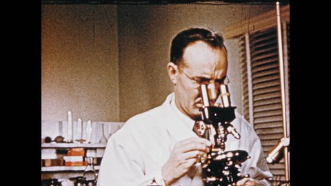 1950s: Man looks through microscope, adjusts magnification power, looks through microscope. Microscopic salt molecules float in water.