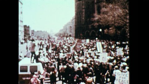 1960s: People march down the street, protest. Men in military uniforms look at protesters. Women stand on steps, hold banner, chant.