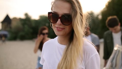 Young pretty woman walking and drinking beer of glass bottle on the beach during sunset, steadycam shot, slow motion. Blonde in glasses quenches thirst with lemonade beverage at sandy sea shore.