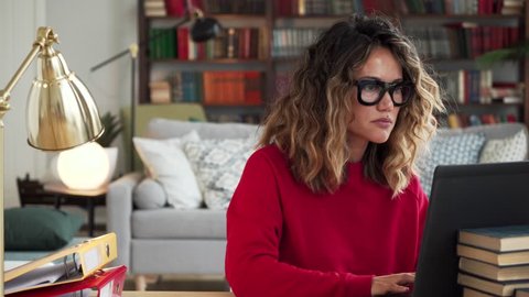 Beautiful curly woman in thirties of fair complexion in red jersey and glasses works on her laptop sitting at the table entering information from a paper into computer in stylish spacious room 库存视频