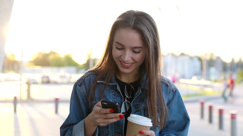 Pretty Young Woman Walking in the City. Using her Mobile Phone. Chatting on it. Typing a Message. Girl Looking Excited, Satisfied. Smiling Happily. Drinking Delicious Coffee. Stylish Outfit. Royalty-Free Stock Footage #1014030491
