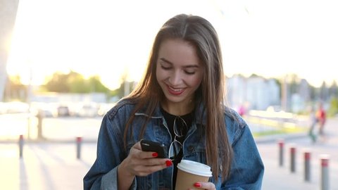 Pretty Young Woman Walking in the City. Using her Mobile Phone. Chatting on it. Typing a Message. Girl Looking Excited, Satisfied. Smiling Happily. Drinking Delicious Coffee. Stylish Outfit.