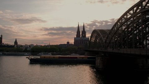 Static clip of Hohenzollernbrucke and Cologne Cathedral at sunset in Germany. A large barge passes under bridge