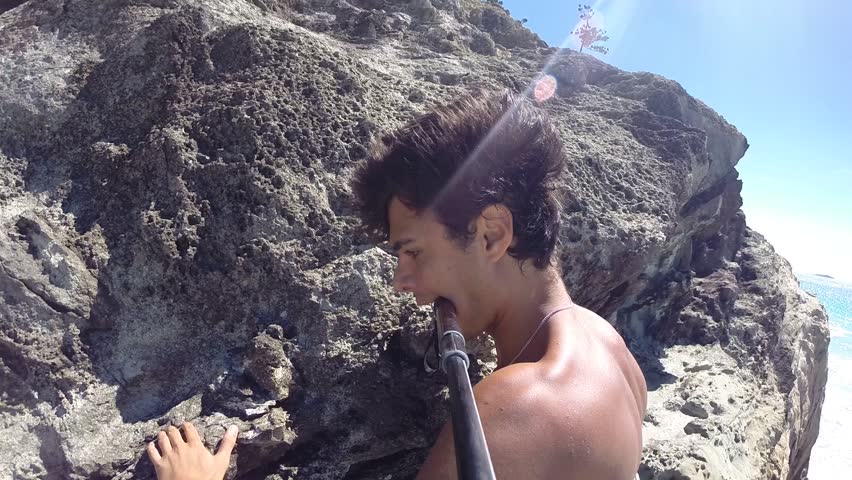 Man holding in his mouth a selfie stick while climbing on rocks along the beach in Australia