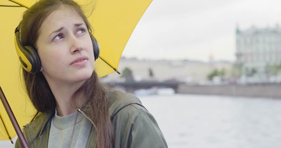 Positive young girl with brown hair and with wireless headphones and smartphone in hands dances on the street of St. Petersburg rejoicing of the spring rain