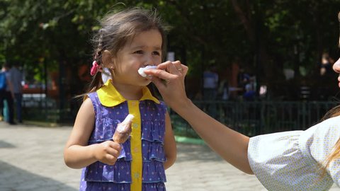 Mom wipes his little daughter's face, soiled with ice cream. In the background a public park.