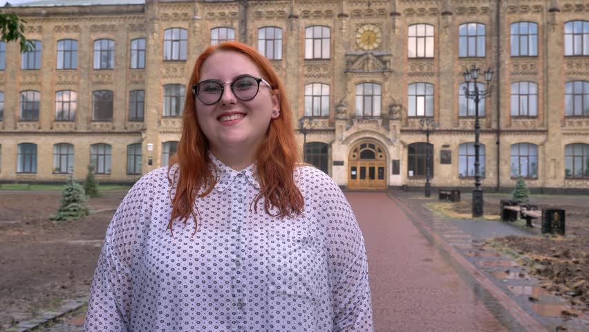 Body positive ginger girl with glasses is standing in park in rainy weather, watching at camera, smiling, building on background