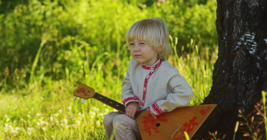 A little boy sits under a tree and plays a balalaika.