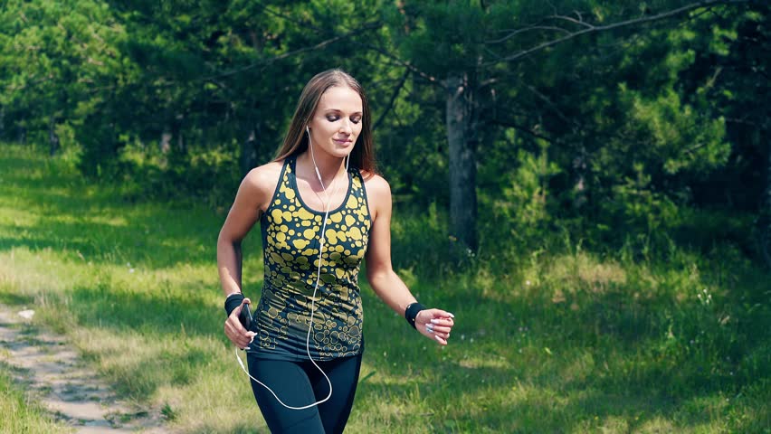 CU, Portrait: Young girl goes in for sports in the morning, runs along a pine forest, she has wristlets on her hands, she listens to music, uses a smartphone, controls the pulse, pressure. 