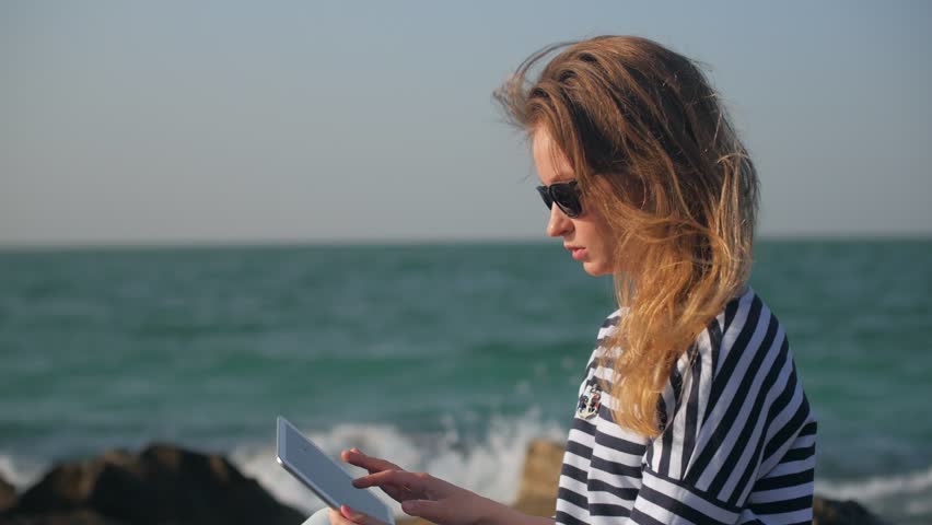 cheerful blonde girl is watching funny photos in her tablet and smiling, sitting outdoors