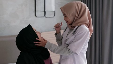 Young asian muslim female doctor with patient. Routine health check and looking at her throat. In doctor exam room. Edited and raw.