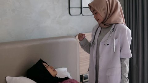 Young asian muslim female doctor in white coat with stethoscope at work. Measuring temperature to sick patient by thermometer at home. Consulting about treatment. Edited and raw.