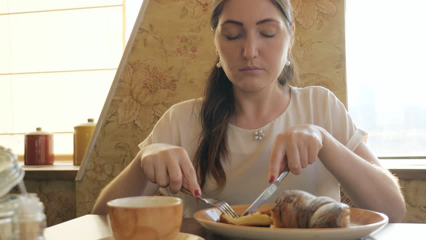 brunette woman eating a croissant and singing coffee at a cafe.