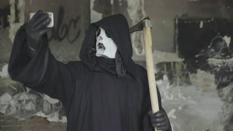 Grim reaper with mower in funny action with cell phone, comic scene with angel of death, dark demon from hell holds mobile and making selfie photos, selfi parody, scary halloween mask and costume.