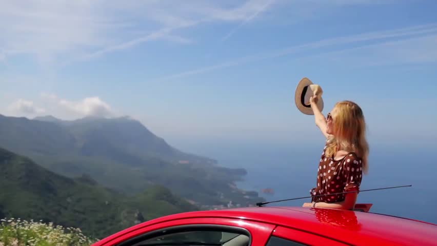 beautiful girl waving her hat from the car with a view of the mountains and the sea