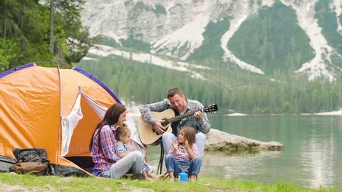 A happy family camping at the lake, playing the guitar and singing a song together in front of the tent. Concept of: trekking, nature, family.
