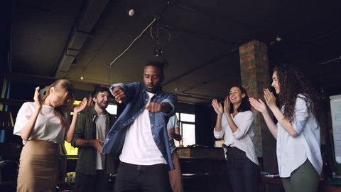 Slow motion of happy office worker African American guy dancing at corporate party while his team members are clapping hands, looking at him and laughing.
