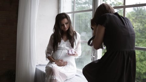 Beautiful young pregnant photo model in white peignoir posing for female photographer while sitting next to the window in bright room