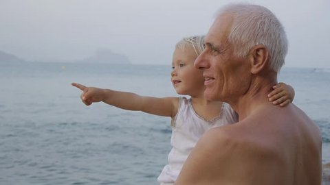 Portrait of senior man holding in arms blonde blue-eyed baby girl on sea coast background. Child pointing to the sea