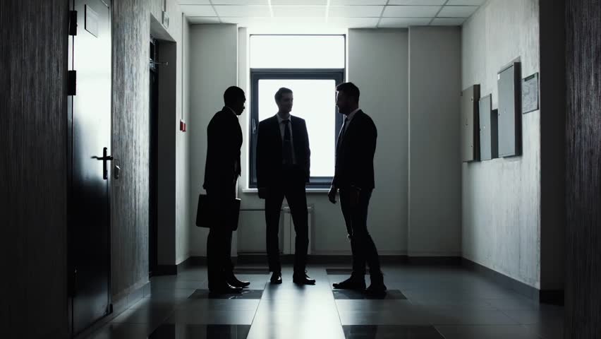 Silhouettes of three businessmen standing in corridor in front of light window. The end of working day. Team completed the work on the project. Businessmen say good-bye and shake hands. Handshake. Royalty-Free Stock Footage #1014121577