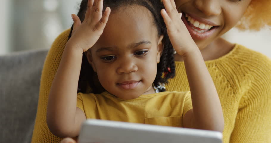 Close up of the little cute African American girl sitting on the knees of her mother, watching something on the screen of tablet and clapping hands. Indoors