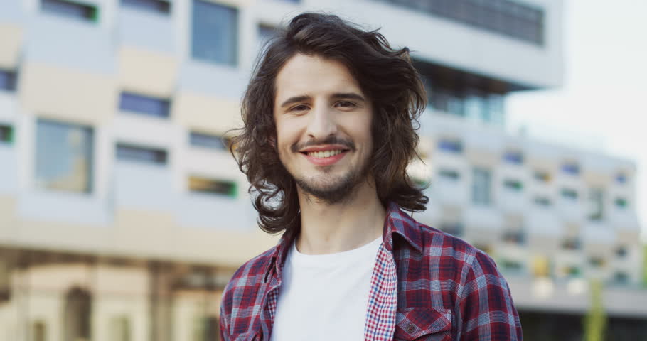 Close up of the young handsome man with long hair in the plaid shirt turning his head to the camera on the urban building background. Outdoor.