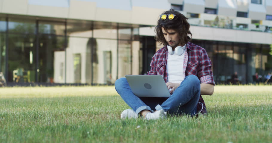Caucasian young man in the motley shirt typing on his laptop computer and sitting on the grass near his college. Outside.