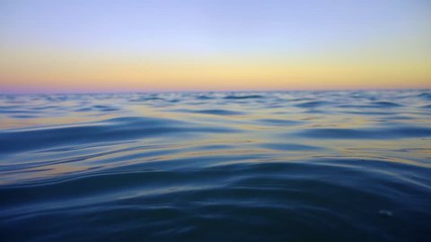 Slow motion rising up from underwater to smooth ocean waves at sunset. Emerging from surface of the sea to orange sky and calm soothing waters. Reaching above water level to gasp air to breathe.