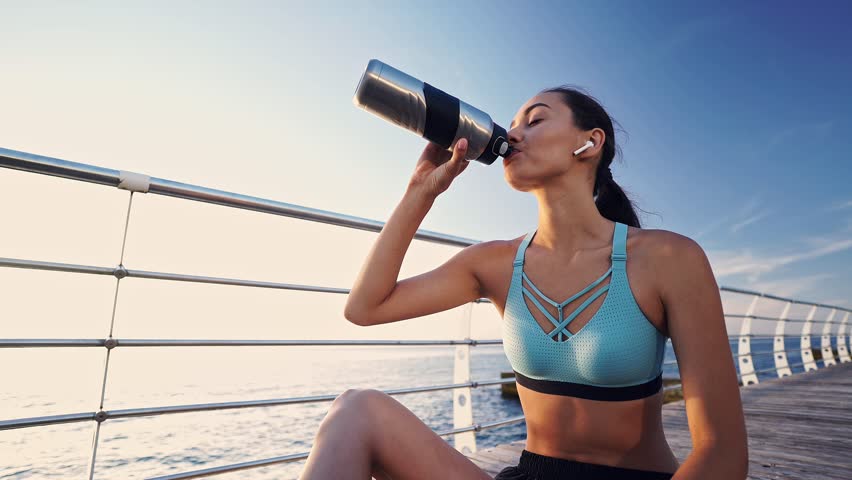 Tired sports woman in earphones sitting near the sea and relax while drinking water