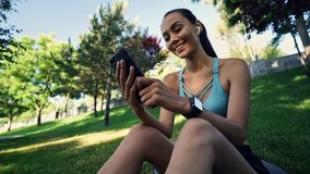 Smiling sports woman in earphones using smartphone while relaxing on grass outdoors
