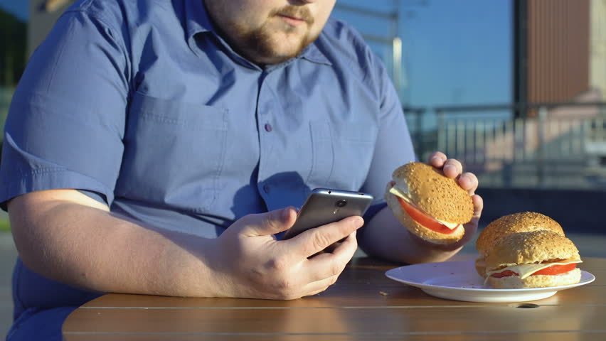 Lazy young man eating unhealthy burger and scrolling smartphone application