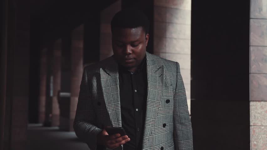Joyful young Afro-American worker in a formal wear walks out of the office, actively uses his phone, and laughs at received messages. Surfing the internet, having fun, break, positive emotions. Male