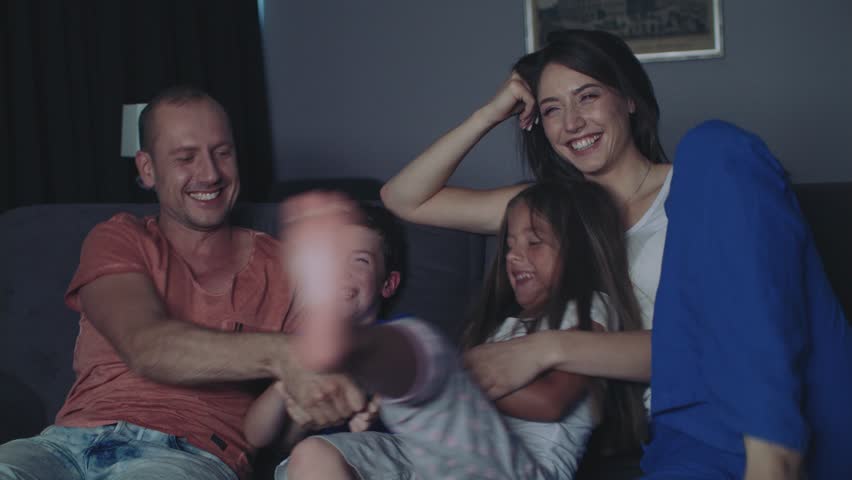 Parents And Their Children Are Watching TV. They Sit On A Sofa In Their Cozy Living Room. It's Evening. Children are fooling and picking up the remote. Royalty-Free Stock Footage #1014156452