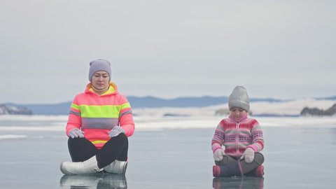 Family is training practice yoga in winter. Woman is do stretching and meditation on ice in nature. Mother and daughter practices yoga on ice in cracks. Girls do sport fitness in outdoor. Background