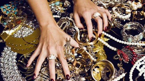 Female hands with a large ring of gold with precious stones touch a bunch of gold and silver jewelry on a black background. Luxurious life. Incredible wealth. Hidden treasures.