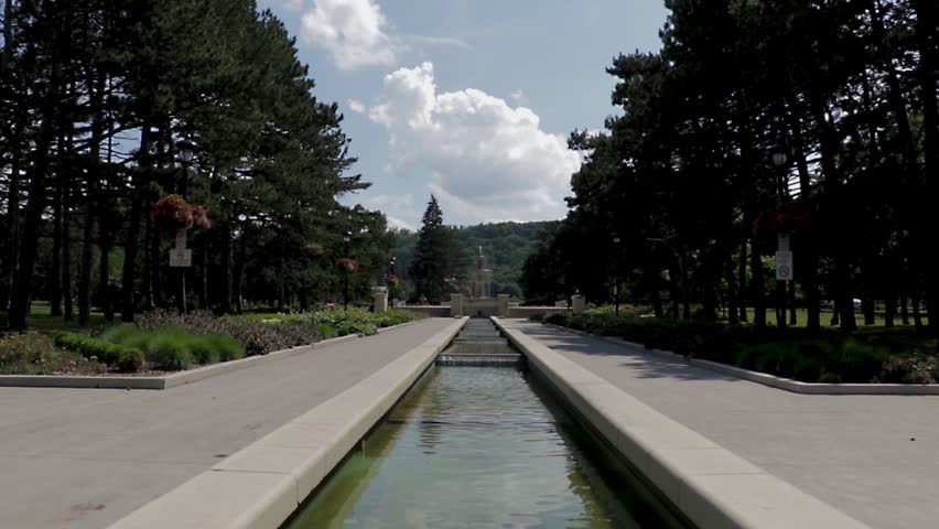 Empty park with large water fountain and trees lining the walkway. Sunny summer day with large white clouds in the sky. Peaceful urban park with nobody around.  
