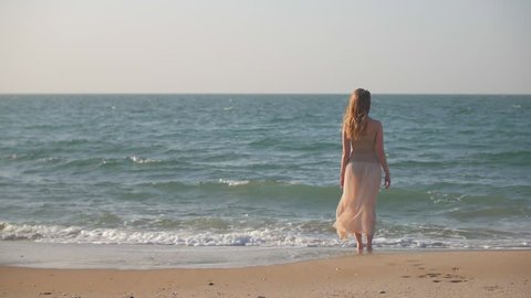 slim blonde girl is standing on a sand beach in seawater and watching waves in summer day