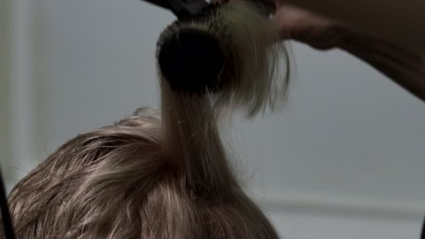 The hairdresser dries blonde hair with a hair dryer and a round hair brush. Closeup.