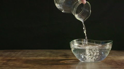 Man pouring water in 180fps slow motion from a clear jug into a curved medium sized bowl on a dark wood table and black background. 