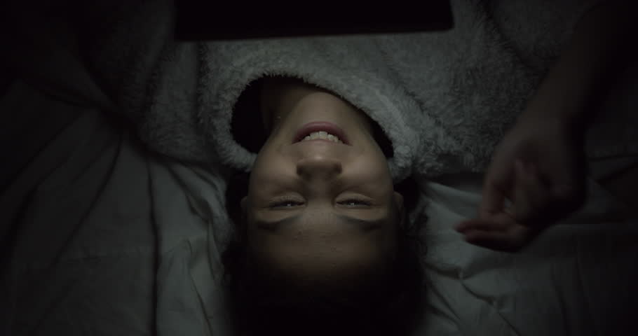 Profile of Mixed Race Teenage Girl on Smart Tablet in Dark Room with Headphones, Student laying with Mobile Night Light, Young Woman Studying Social Media on Bed