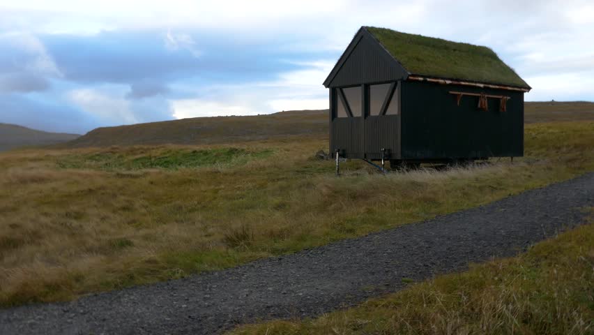 Nordic style wooden house with grass roof and reflection of sky and clouds on the window located on hill near a pathway