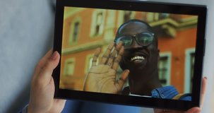 Female hands waving while holding a tablet computer with an attractive smiled African American man in glasses videochatting on its screen. Close up.