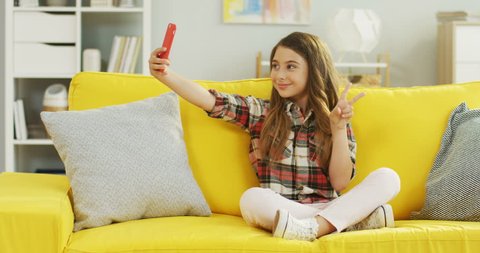 charming girl taking selfie photos on smartphone device and making gestures on sofa at home. Indoor.